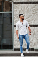 Fototapeta na wymiar Stylish tall arabian man model in white shirt, jeans and sunglasses posed at street of city. Beard attractive arab guy.