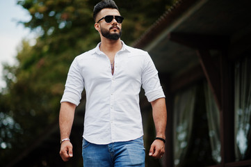 Stylish tall arabian man model in white shirt, jeans and sunglasses walking at street of city....
