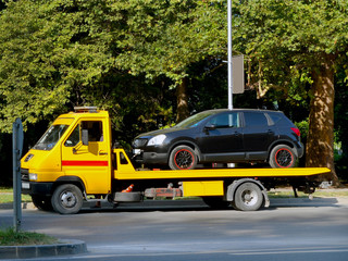 Black car is loaded on a yellow car tow truck on a city street on a summer day