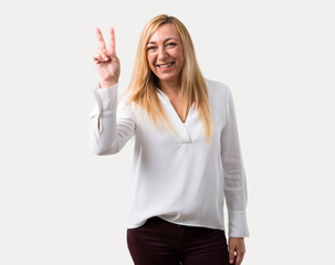 Fototapeta premium Middle age blonde woman with white shirt smiling and showing victory sign with a cheerful face on isolated grey background