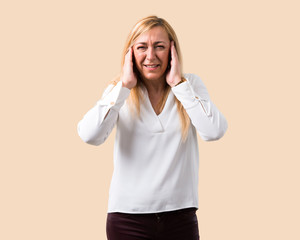 Middle age blonde woman with white shirt covering both ears with hands. Frustrated expression on isolated ocher background