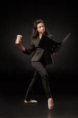attractive businesswoman in suit and ballet shoes with folder and coffee to go posing on black backdrop