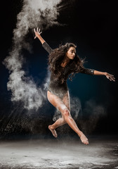 Fototapeta na wymiar beautiful ballerina in black bodysuit jumping on dark background with talc powder around