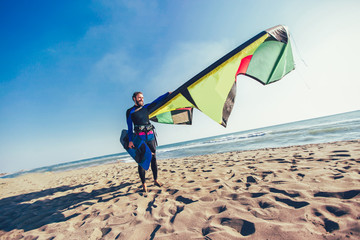 Handsome Caucasian man professional surfer standing  on the sandy beach with his kite and board.