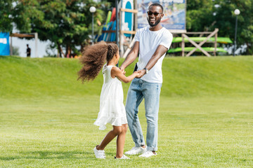 Fototapeta premium happy african american father and daughter holding hands and spinning around in amusement park
