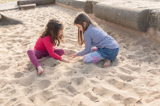 Little Girls With Bare Feet Sitting And Playing Together In Sand Pit At Fenced Playground With Afternoon Shadows (selective Focus)