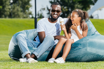 smiling african american father and daughter with ice cream having fun on beanbag chairs in park