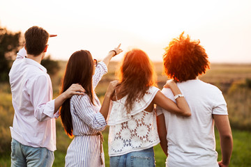 Two young guys and two girls dressed in a stylish clothes are standing in the field and looking in front of them on a sunny day.