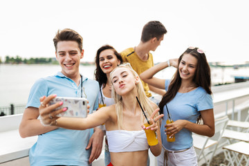 A company of good-looking friends laughing and drinking yellow cocktails and socialising and making selfie in the nice cafe next to the river. Cheers. Entertainment, having good time. 