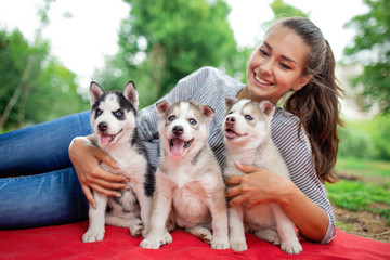 A beautiful smiling woman with a ponytail and wearing a striped shirt is cuddling with  three sweet husky puppies while resting on the red blanket on the lawn. Love and care for pets.