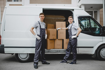 Two young handsome smiling workers wearing uniforms are standing next to the van full of boxes. House move, mover service. - obrazy, fototapety, plakaty