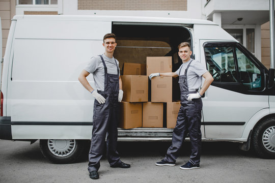 Two young handsome smiling workers wearing uniforms are standing next to the van full of boxes. House move, mover service.