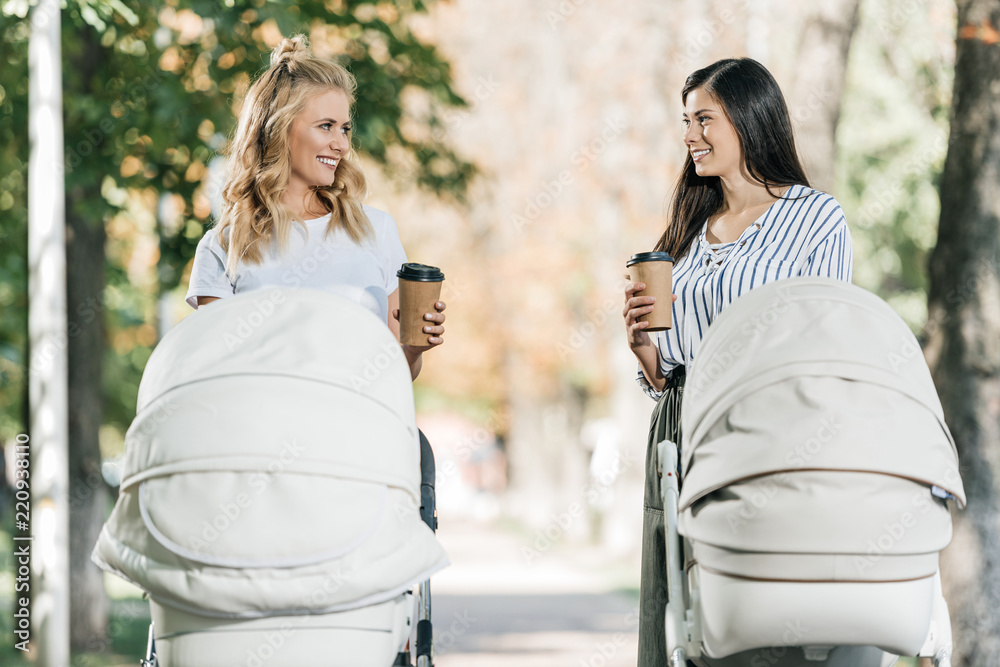 Wall mural happy mothers walking with baby strollers and coffee in paper cups in park