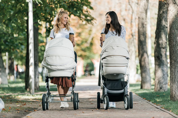 mothers walking with baby strollers and coffee to go in park and looking at each other