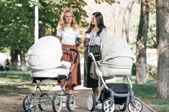 smiling mothers holding coffee to go and walking with baby strollers in park