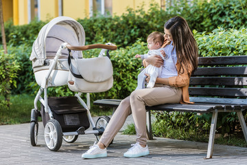 mother sitting with baby on bench near stroller in park