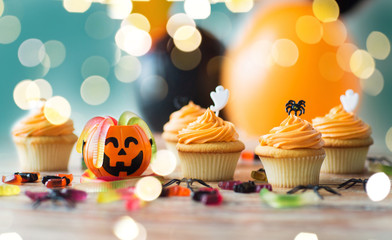 food, baking and holidays concept - cupcakes or muffins and candies with halloween party decorations on wooden table over lights