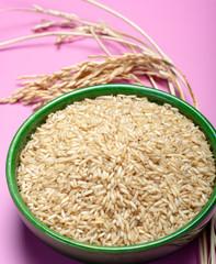 Bowl with organic brown uncooked rice and dried rice plant on pink background