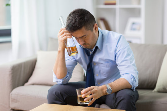 Alcoholism, Alcohol Addiction And People Concept - Male Alcoholic With Bottle And Glass Drinking Whiskey At Home