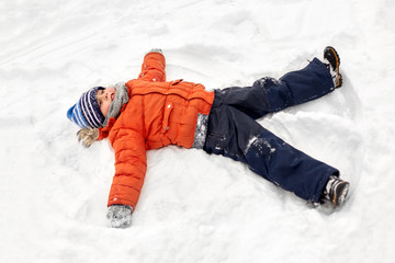 Fototapeta na wymiar childhood, leisure and season concept - happy little boy in winter clothes making snow angels outdoors