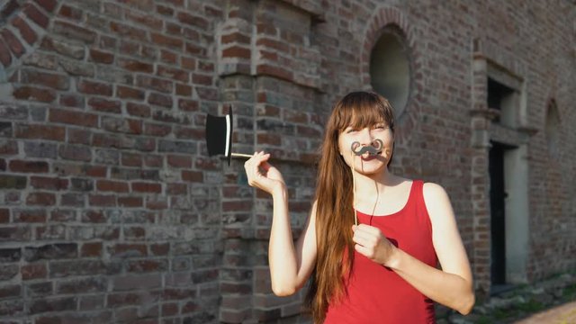 Young happy woman holds a paper mustache and black retro hat on a stick, dancing next to a brick wall.