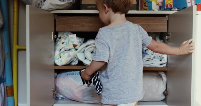 Kid opens the chest of the chest of drawers. Blond toddler tries to open door of locker back view