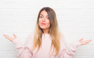 Young adult woman over white brick wall wearing winter outfit at home clueless and confused expression with arms and hands raised. Doubt concept.