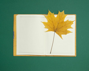 Open blank notebook with a yellow autumn maple leaf on dark green background top view flat lay. Autumn concept, study, working table, workspace mockup.