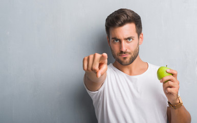 Handsome young man over grey grunge wall eating green apple pointing with finger to the camera and to you, hand sign, positive and confident gesture from the front