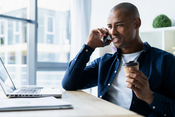 Young man talking on his mobile phone in office. African executive sitting at his desk with laptop