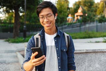 Happy asian male student in eyeglasses and earphones using smartphone