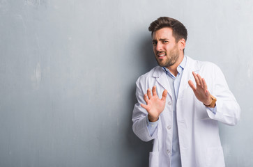 Handsome young professional man over grey grunge wall wearing white coat disgusted expression, displeased and fearful doing disgust face because aversion reaction. With hands raised. Annoying concept.