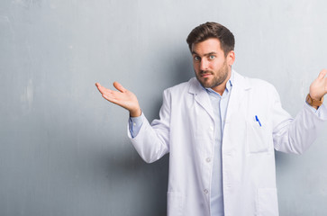 Handsome young professional man over grey grunge wall wearing white coat clueless and confused expression with arms and hands raised. Doubt concept.