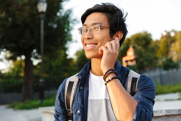 Smiling asian male student in eyeglasses talking by smartphone