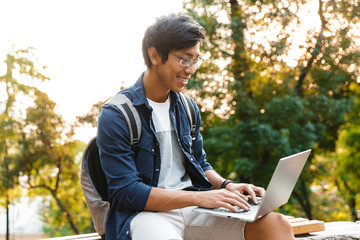 Pleased asian male student in eyeglasses using laptop computer
