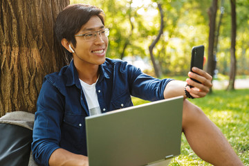 Happy asian male student in eyeglasses making selfie on smartphone