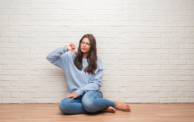 Young brunette woman sitting on the floor over white brick wall with angry face, negative sign showing dislike with thumbs down, rejection concept