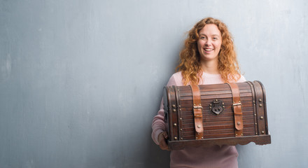 Young redhead woman holding vintage chest with a happy face standing and smiling with a confident...