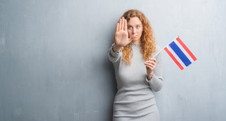 Young redhead woman over grey grunge wall holding flag of Thailand with open hand doing stop sign...