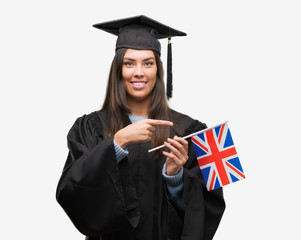 Young hispanic woman wearing graduated uniform holding flag of united kingdom very happy pointing...