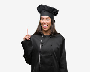 Young hispanic cook woman wearing chef uniform surprised with an idea or question pointing finger with happy face, number one