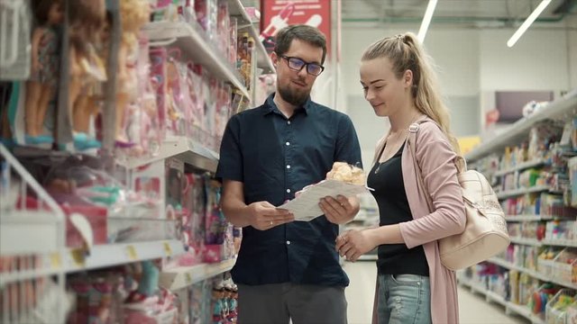 Man and woman are choosing gift for their children in a store. They are standing in toy department near racks, mother is holding dolls in hands