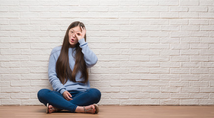 Young Chinese woman sitting on the floor over brick wall doing ok gesture shocked with surprised face, eye looking through fingers. Unbelieving expression.