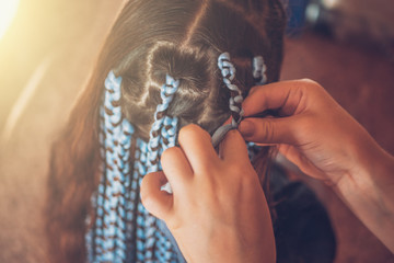 Hairdresser weaves braids with kanekalon material to young girl head, making creative hairstyle with thick plaits or pigtails also known as Afro braids - obrazy, fototapety, plakaty