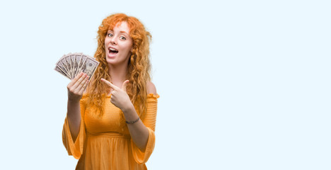 Young redhead woman holding dollars very happy pointing with hand and finger