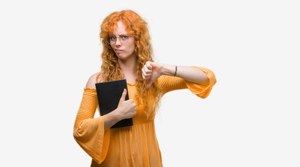 Young redhead student woman holding a book with angry face, negative sign showing dislike with thumbs down, rejection concept