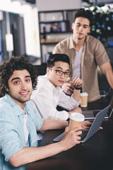 three multiethnic businessmen with paper coffee cups sitting at table with laptops at modern office