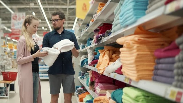 Two cheerful people are discussing buying of textile for home. Wife and husband is touching different kind of towels in a supermarket