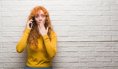 Young redhead woman standing over brick wall talking on the phone cover mouth with hand shocked with shame for mistake, expression of fear, scared in silence, secret concept