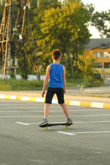 Obraz premium Boy riding skate board on summer parking lot near power station, back view, sunset